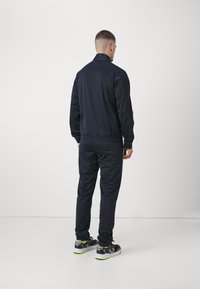 Tuta blu navy con colletto alto, polsini ed elastico in vita. Il materiale appare liscio, abbinato a sneaker nere con dettagli neon.