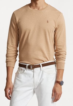 Beżowa koszulka z długim rękawem o okrągłym dekolcie, dopasowanym kroju i małym haftowanym logo na piersi; zestawiona z białymi spodniami i brązowym paskiem.