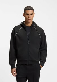 Mann trägt schwarzen Kapuzenpullover mit Reißverschluss und weißer Paspelierung sowie schwarze Hose, steht vor einem schlichten weißen Hintergrund.