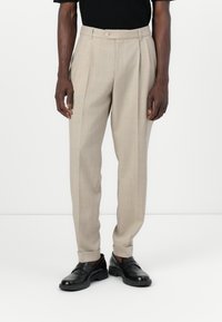 Pantalons plissés beiges avec une coupe ajustée, fabriqués à partir d'un tissu texturé. Présente une fermeture à un bouton et des ourlets à revers. Associés à des chaussures noires.