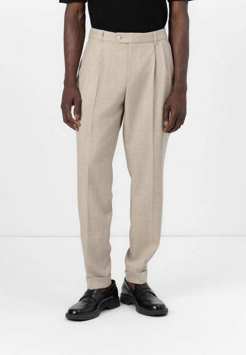 Pantalons plissés beiges avec une coupe ajustée, fabriqués à partir d'un tissu texturé. Présente une fermeture à un bouton et des ourlets à revers. Associés à des chaussures noires.