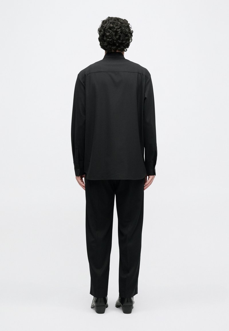 Schwarzes Langarmshirt mit geradem Schnitt, weichem Stoff, Naht am Rückenpasse, kombiniert mit passenden locker sitzenden Hosen und schwarzen Schuhen.