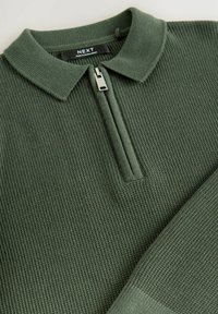 Pullover lavorato a maglia verde scuro con colletto a zip, caratterizzato da una texture a coste e un'etichetta con la scritta "NEXT". Design semplice e pulito con una zip frontale.