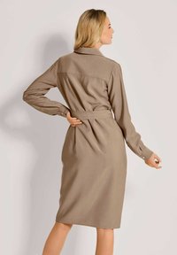 Lange mouwen, geknoopte jurk van lichtbeige stof. Heeft een getailleerde taille met een riem en knoopmanchetten. Glad textuur.