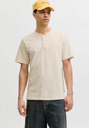 Uomo con maglietta henley beige a maniche corte, jeans scuri e un cappellino giallo, in piedi davanti a uno sfondo bianco uniforme.