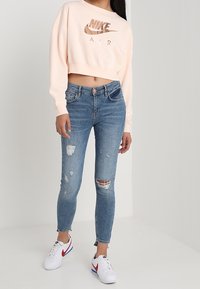 Světle růžový zkrácený svetr s logem "Nike Air", spárovaný s roztrhanými modrými skinny džíny. Bílé tenisky s červenými a modrými akcenty.