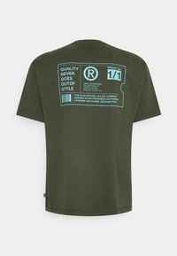 Olivově zelené bavlněné tričko s krátkým rukávem. Na zadní straně má modrý grafický štítek s designem a textem a ochrannou známku.