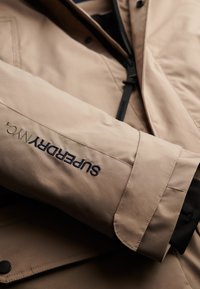 Béžová nepromokavá bunda s hladkou texturou, se zpevněným černým zipem a švy. Logo "SUPERDRY" je vyšité na rukávu.
