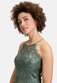 Zielona sukienka koronkowa z misternym haftem kwiatowym, przezroczystą warstwą i kreacją na szyi z koralikowymi akcentami. Tekstura wydaje się gładka.