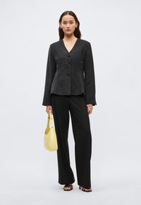 Čierny štruktúrovaný blejzer s výstrihom do V, obsahujúci päť gombíkov a dlhé rukávy, v kombinácii s čiernymi širokými nohavicami a žltou kabelkou.