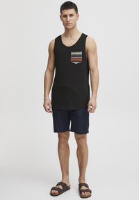 Top negru fără mâneci cu un buzunar frontal cu model, asortat cu pantaloni scurți bleumarin și sandale maro. Topul are o croială lejeră și o textură moale.