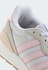 Bijela tenisica s ružičastim kožnim prugama i bež antilop detaljima. Ima mrežasti gornji dio, teksturiranu površinu i bijeli jastučićasti rub.