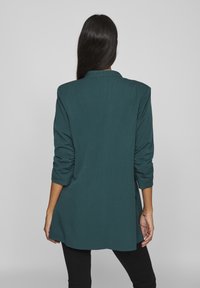 Tyrkysový blazer se strukturovaným designem, s ohrnutými rukávy a přizpůsobenou siluetou. Hladká textura s mírným leskem.