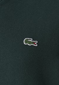 Pullover verde in maglia con un piccolo logo di un coccodrillo ricamato in bianco e rosso sul lato sinistro del petto. Il materiale sembra liscio ed elastico.