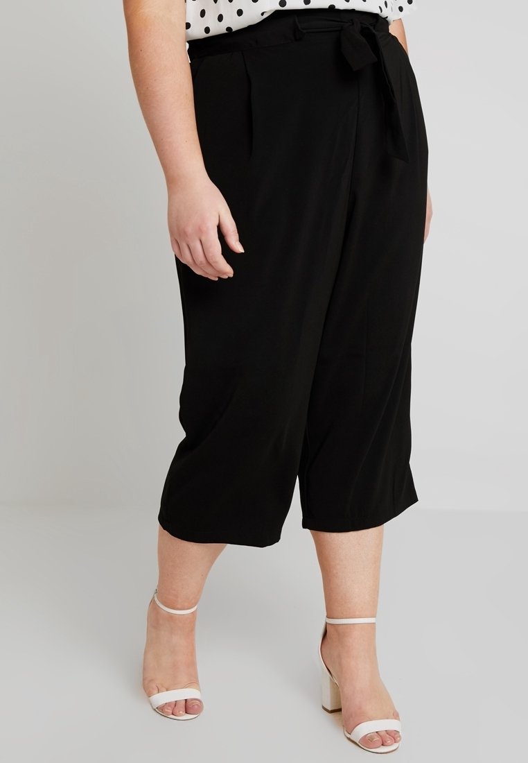 Czarne krótkie spodnie z wiązaniem w talii, o gładkiej fakturze i bocznymi kieszeniami, zestawione z białymi sandałkami na obcasie oraz bluzką w kropki.