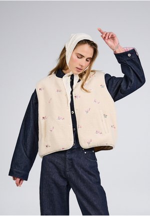 Młoda kobieta ubrana w kremową, kwiatową kamizelkę z polaru na ciemnej dżinsowej koszuli i dżinsach, z białą chustą zawiązaną pod brodą.