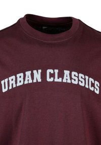 Bordo krūšturis ar garām piedurknēm, izgatavots no kokvilnas, ar baltu uzrakstu "URBAN CLASSICS", kas ar izliekumu izvietots pār krūtīm treknās burtiem.