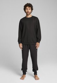 Schwarzes Langarm-Sweatshirt mit Rundhalsausschnitt, kombiniert mit strukturierten schwarzen Jogginghosen mit kleinem Logo. Beides ist eng geschnitten und lässig.