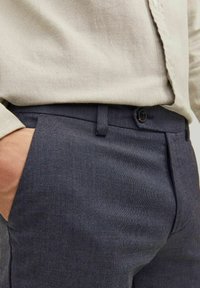 Laivastonsiniset tekstuurihousut, joissa on hentoa kuviointia, nappikiinnitys ja vyölenkit, yhdistettynä vaaleanbeigeen paitaan.