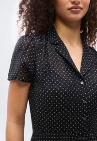 Γυναίκα που φοράει μαύρο διαφανές πουκάμισο με λευκές πουά και κοντά μανίκια φουσκωτά, κουμπωμένο μπροστά.