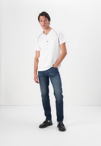 Jonge man in een aansluitend witte V-hals shirt en blauwe jeans staat casual met zijn handen in de zakken tegen een plain witte achtergrond.