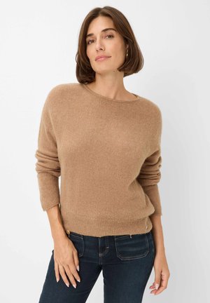 Pull en laine brun clair avec une coupe décontractée, encolure bateau et manches retroussées, associé à un jean foncé. Texture lisse avec une douceur subtile.