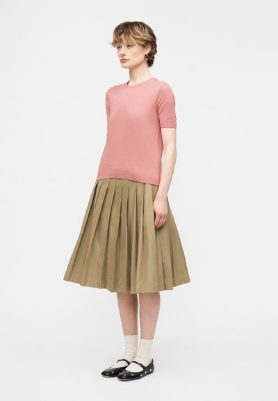 Γυναίκα που στέκεται, φοράει ροζ μπλούζα με κοντά μανίκια, φούστα μίντι με πιέτες σε ελιά πράσινο, λευκές κάλτσες και μαύρα παπούτσια Mary Jane με ασημένιες θηλιές.