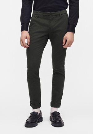 Homme portant un pantalon slim vert foncé retroussé aux chevilles, des chaussettes blanches, des mocassins noirs et une chemise noire à manches longues, debout devant un fond blanc.
