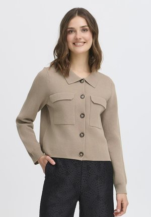 Cardigan en tricot beige avec un col, fermeture à boutons sur le devant et deux poches poitrine ; porté sur un pantalon noir texturé.