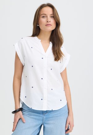 Jeune femme portant un chemisier blanc à manches courtes avec de petites fleurs bleues et un jean bleu clair, debout devant un fond clair uni.