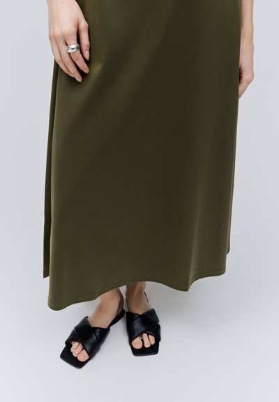 Personne portant une robe longue olive jusqu'aux chevilles et des sandales noires à lanières croisées et orteils ouverts, debout sur une surface claire.