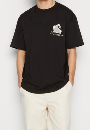 Personne portant un T-shirt noir avec un petit motif blanc et le texte « Carhartt International Pest Control » sur la poitrine gauche et un pantalon beige clair.