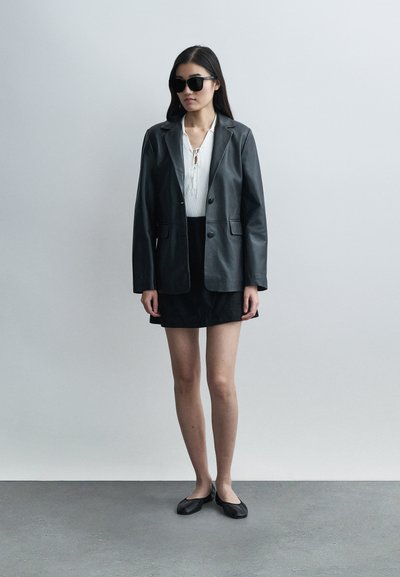 Black leather blazer with notched lapels, worn over a white textured top, paired with a black skirt and flat black shoes. Oversized sunglasses worn.