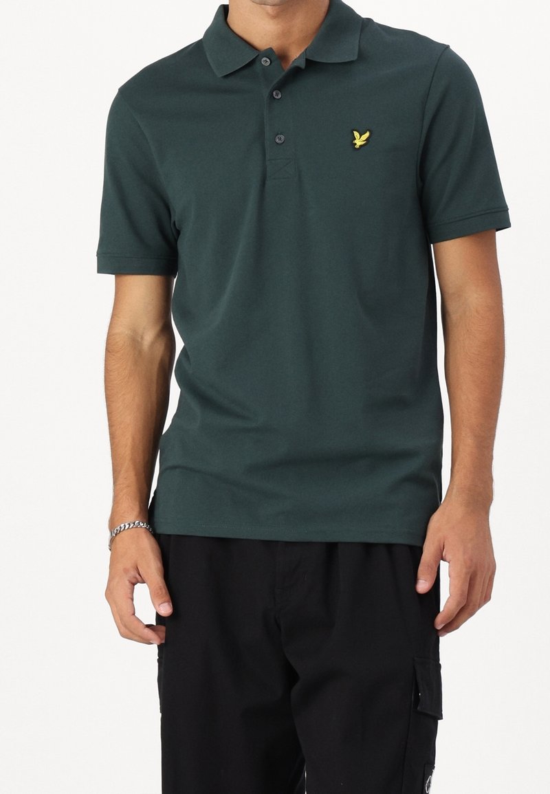 Polo vert foncé avec col, trois boutons et un accent de logo jaune. Confectionné dans un tissu lisse et respirant, silhouette ajustée.
