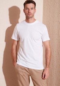 T-shirt bianco a maniche corte realizzato in cotone, con scollo rotondo e vestibilità morbida. Il modello è posizionato contro un muro beige testurizzato.