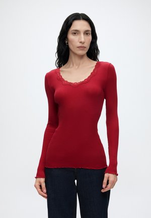Femme aux longs cheveux noirs portant un haut rouge ajusté à manches longues avec bordure en dentelle et un jean bleu foncé, debout devant un fond uni.