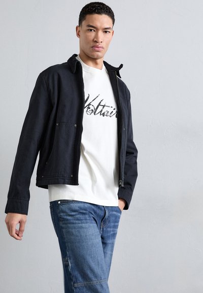 Dark blue zip-up jacket with a collar, paired with a white t-shirt featuring black graphic text; blue denim jeans. Smooth texture, casual design.