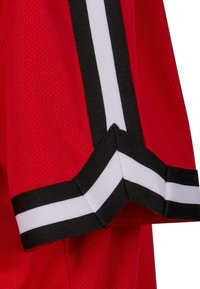 Țesătură de tricou sport roșu cu textură de plasă, având detalii cu dungi negre și albe pe mâneci și un detaliu în formă de V la tiv.