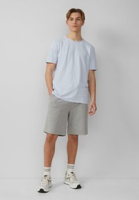 Lichtblauwe katoenen T-shirt met korte mouwen, gecombineerd met grijze katoenen shorts. Model draagt beige sneakers met lichte strepen.
