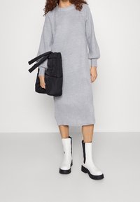 Robe en tricot côtelé grise avec des manches longues, longueur genoux. Sac noir rembourré avec des sangles et bottines blanches avec des détails noirs sur les côtés.