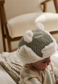 Zelený manšestrový klobúk s bielymi huňatými doplnkami a detailmi pripomínajúcimi uši. Dizajn obsahuje vertikálne rebrá a mäkký, textúrovaný okraj.