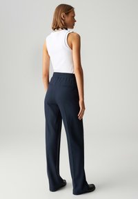 Marineblauwe tapered broeken met een elastische tailleband en twee achterzakken, gecombineerd met een witte mouwloze top. Gladde stoftextuur, minimalistisch ontwerp.