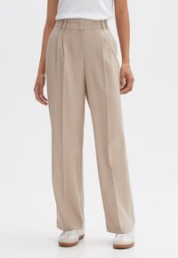 Beige high-waisted wijde broek met plooien, gedragen met witte sneakers en een ingestopt wit topje door een staande persoon.