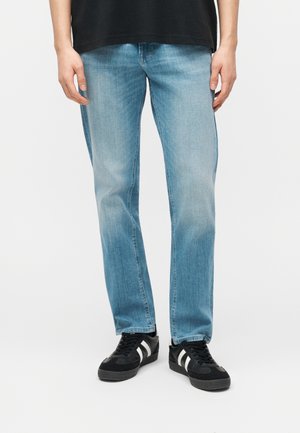 Homme portant un jean droit bleu clair et des baskets noires à rayures blanches, debout devant un fond blanc uni.