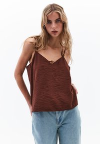 Camisola marrone con texture e scollatura a V, caratterizzata da spalline sottili e un bottone decorativo al centro. Indossata con jeans azzurri chiari.