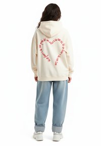 Felpa color beige chiaro con un grande design di testo a forma di cuore rosso sul retro. Abbinata a jeans larghi blu chiaro e sneaker bianche.