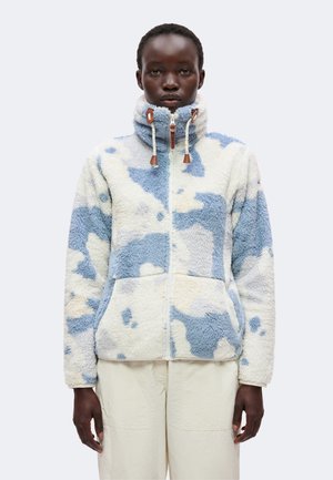 Person trägt eine Fleecejacke mit hohem Kragen mit weißen und blauen abstrakten Mustern und Fronttaschen, kombiniert mit hellen Hosen.
