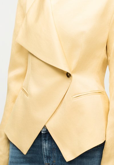 Ανοιχτό μπεζ ασύμμετρο σακάκι με ένα σκοτεινό κουμπί για κούμπωμα, γιακά με πτερύγιο και δύο εμπρόσθιες τσέπες με σχισμή, φορεμένο πάνω από μπλε τζιν.