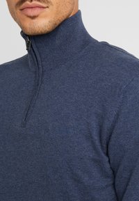 Mörkblå zip-neck tröja i mjukt stickat material, med en ribbad krage och en liten logotyp på bröstet, som visar en figursydd design.