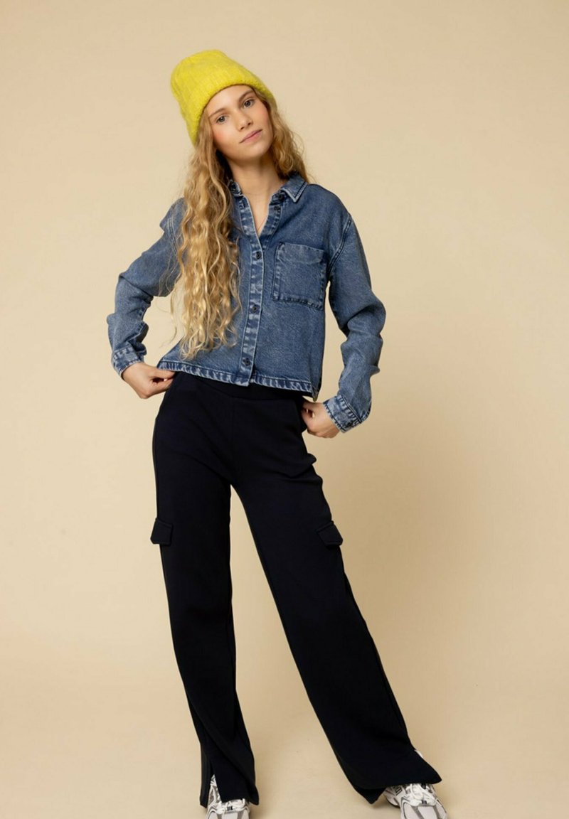 Blauwe spijkerjack met een cropped snit, gecombineerd met zwarte wijde cargobroek met zijzakken. Gele gebreide beanie op het model.